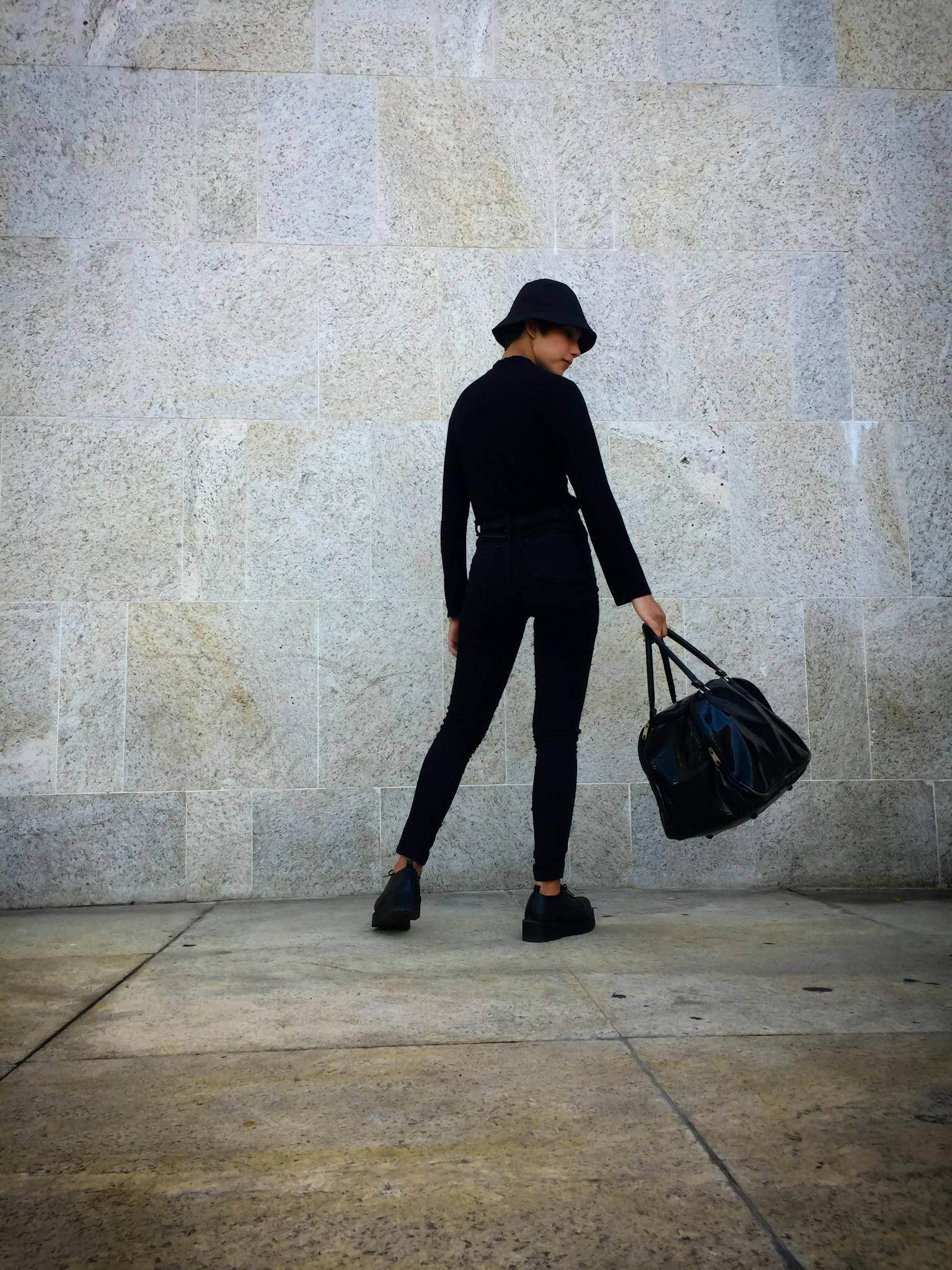 Woman in black outfit and hat poses with a bag against a stone wall. Urban street fashion.